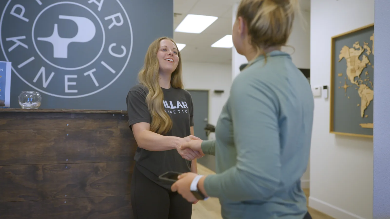 A therapist greeting a patient at reception, in front of the Pillar Kinetic logo