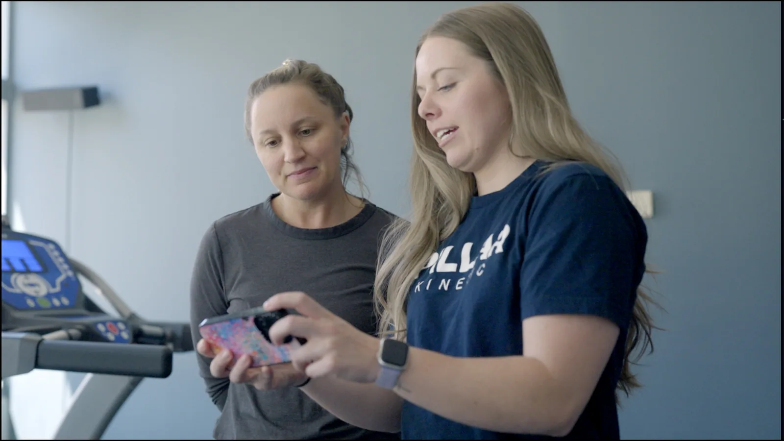 A physical therapist and a patient reviewing a video on a phone beside a treadmill